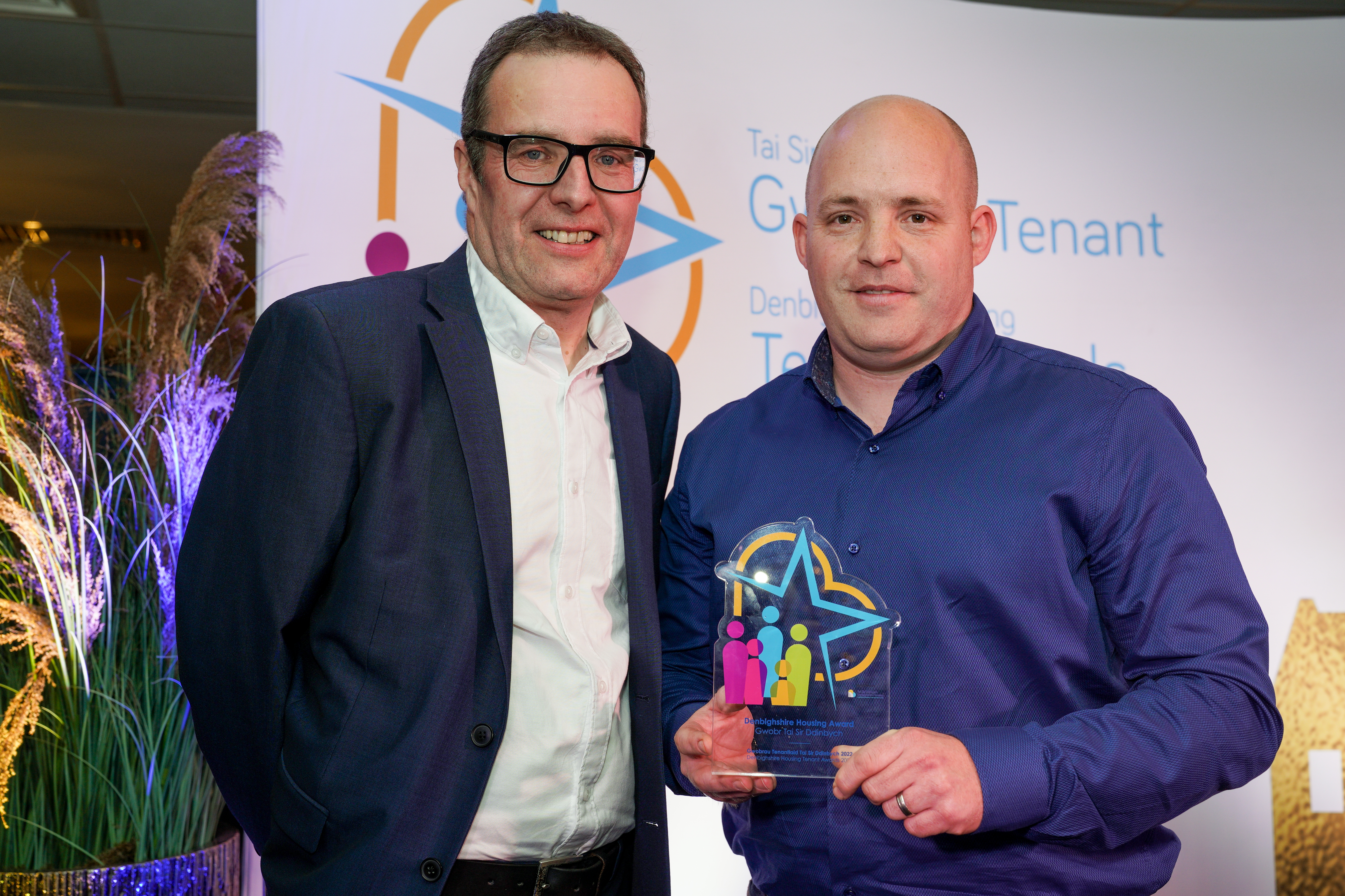 Two men stand together at an award event, with the man in a blue shirt holding a colourful trophy. 