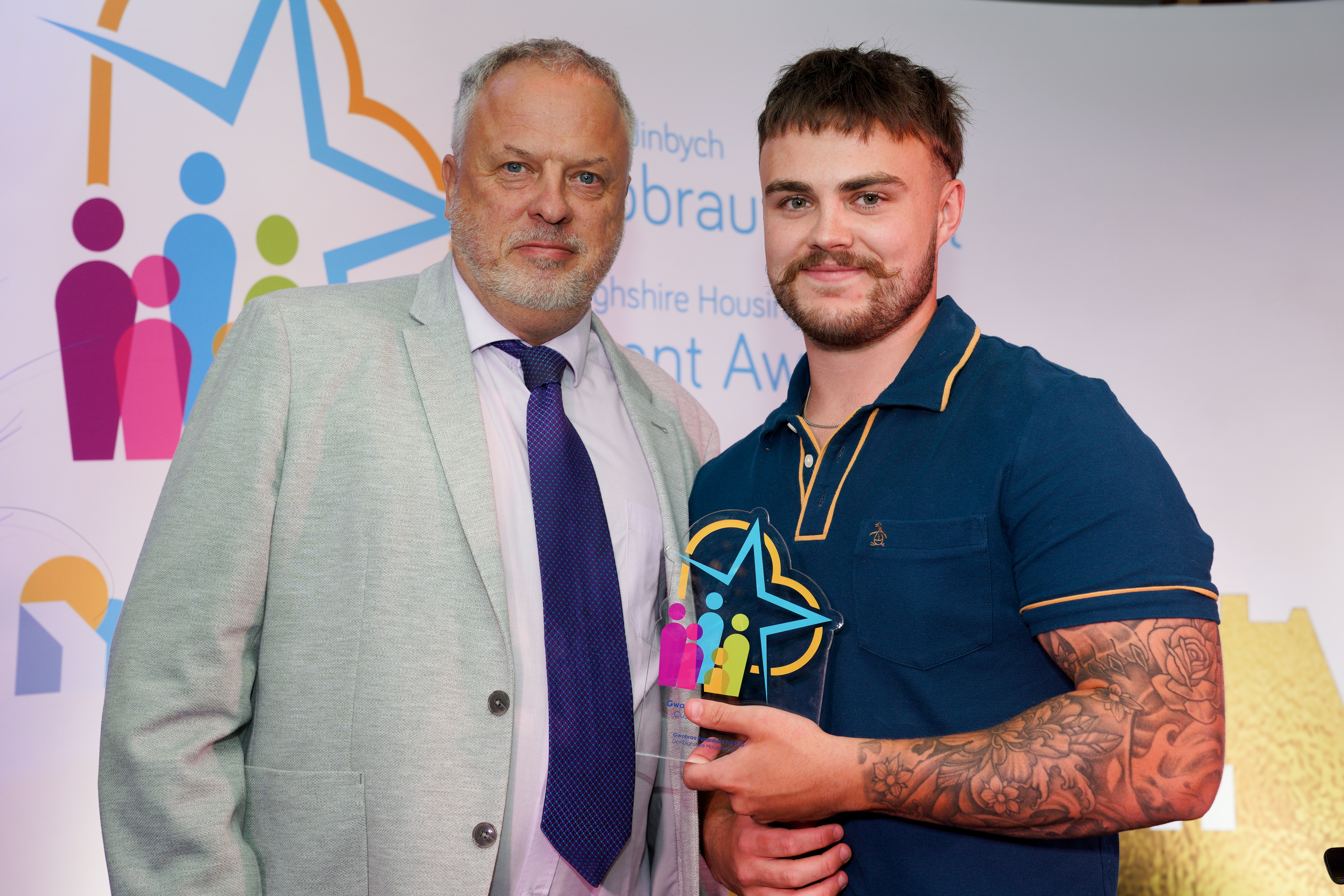 Two men stand together at an award event, with the younger man holding a colourful trophy.