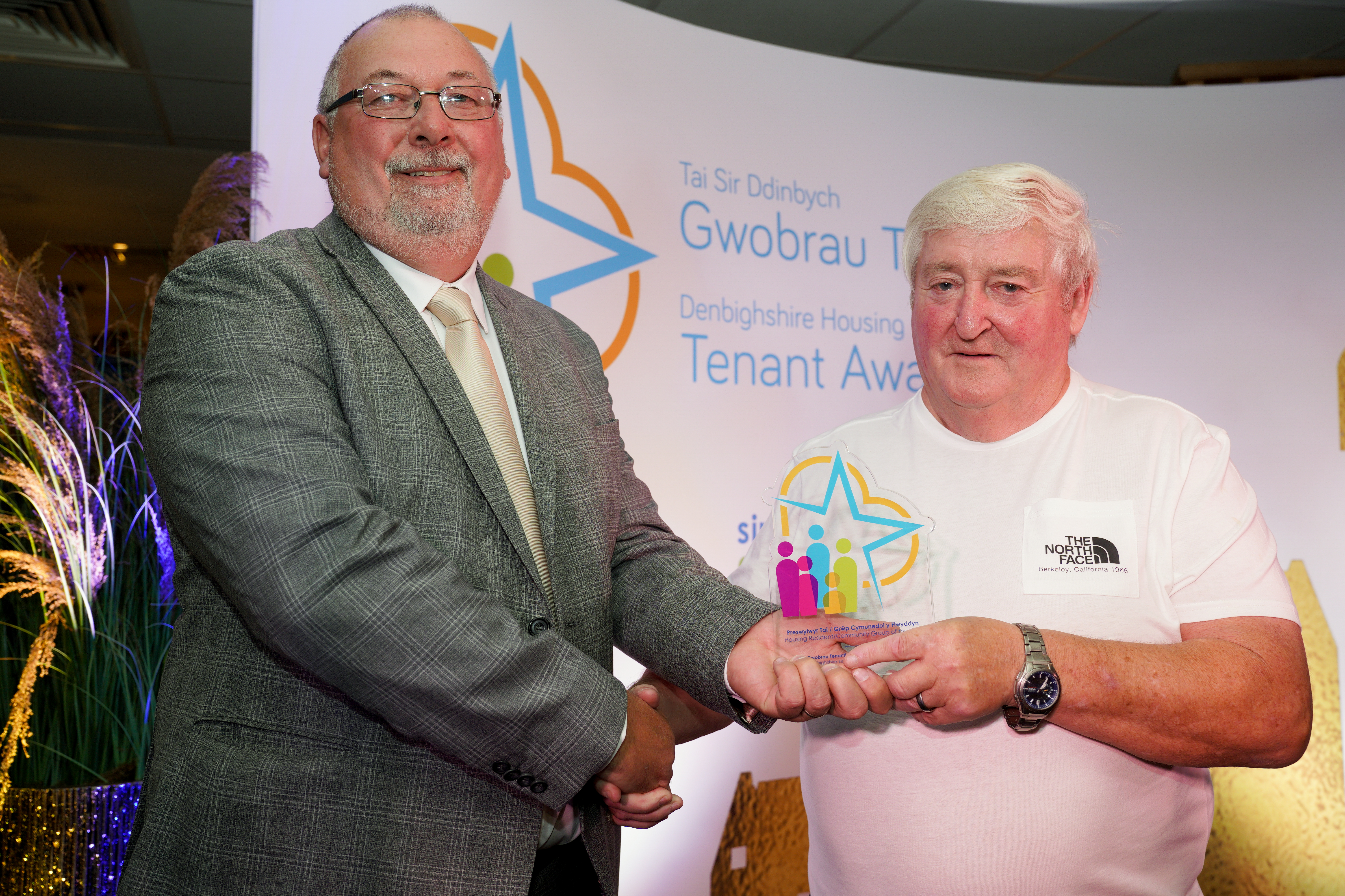 Two men stand together at an award event. A man in a grey suit hands a colourful trophy to a man wearing a white t-shirt.