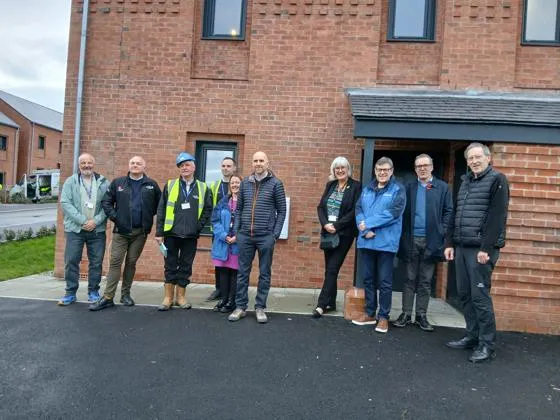 Ten people standing outside a red brick two storey house, wearing business attire.