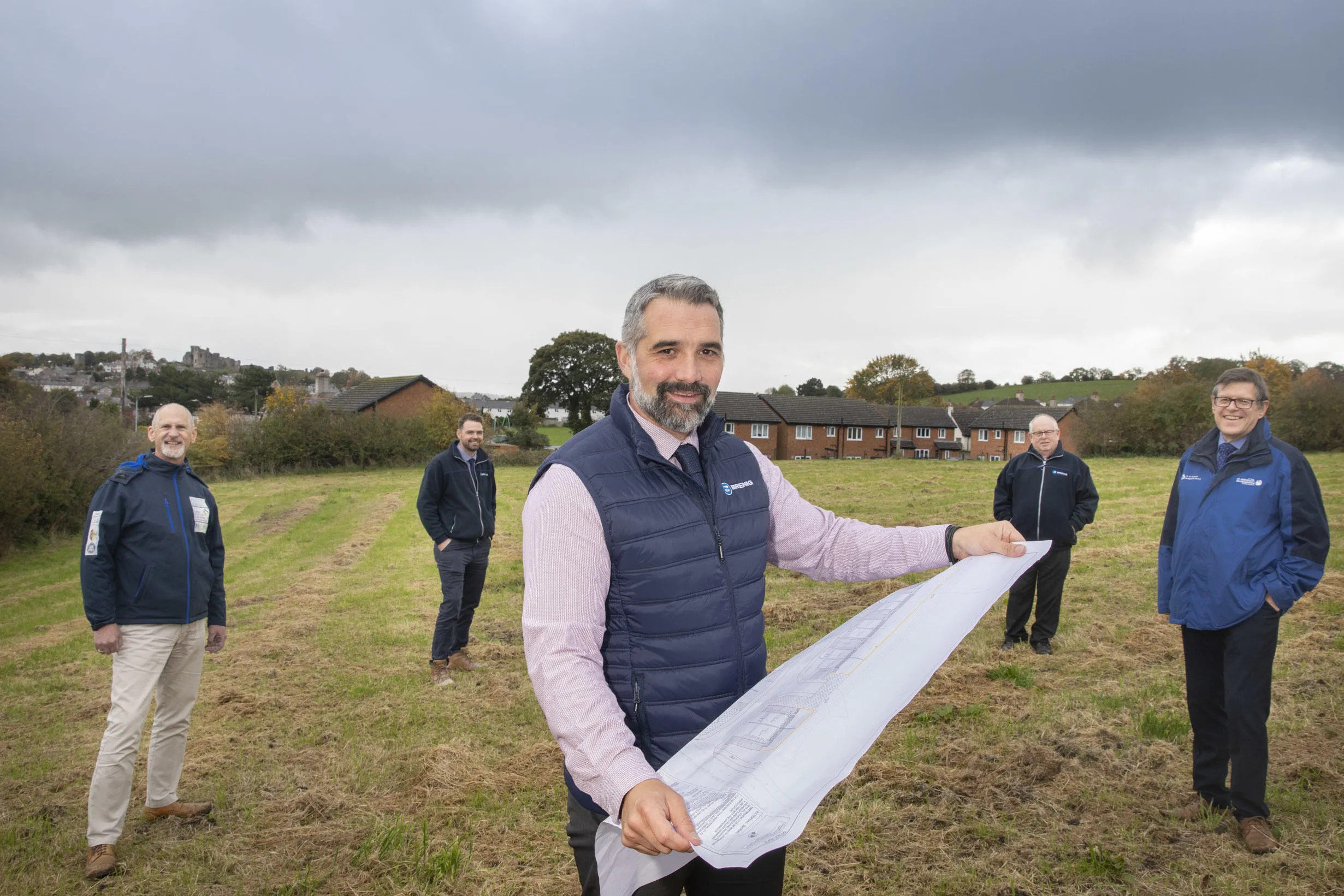 Five men stand together on an area of open land with a housing development plan