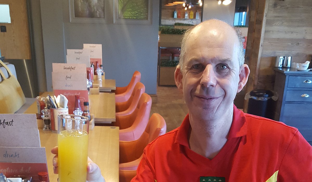 A man in a red shirt sits at a restaurant table, smiling and holding a glass of orange juice. The restaurant has orange chairs, wooden decor, and breakfast menus on the tables.