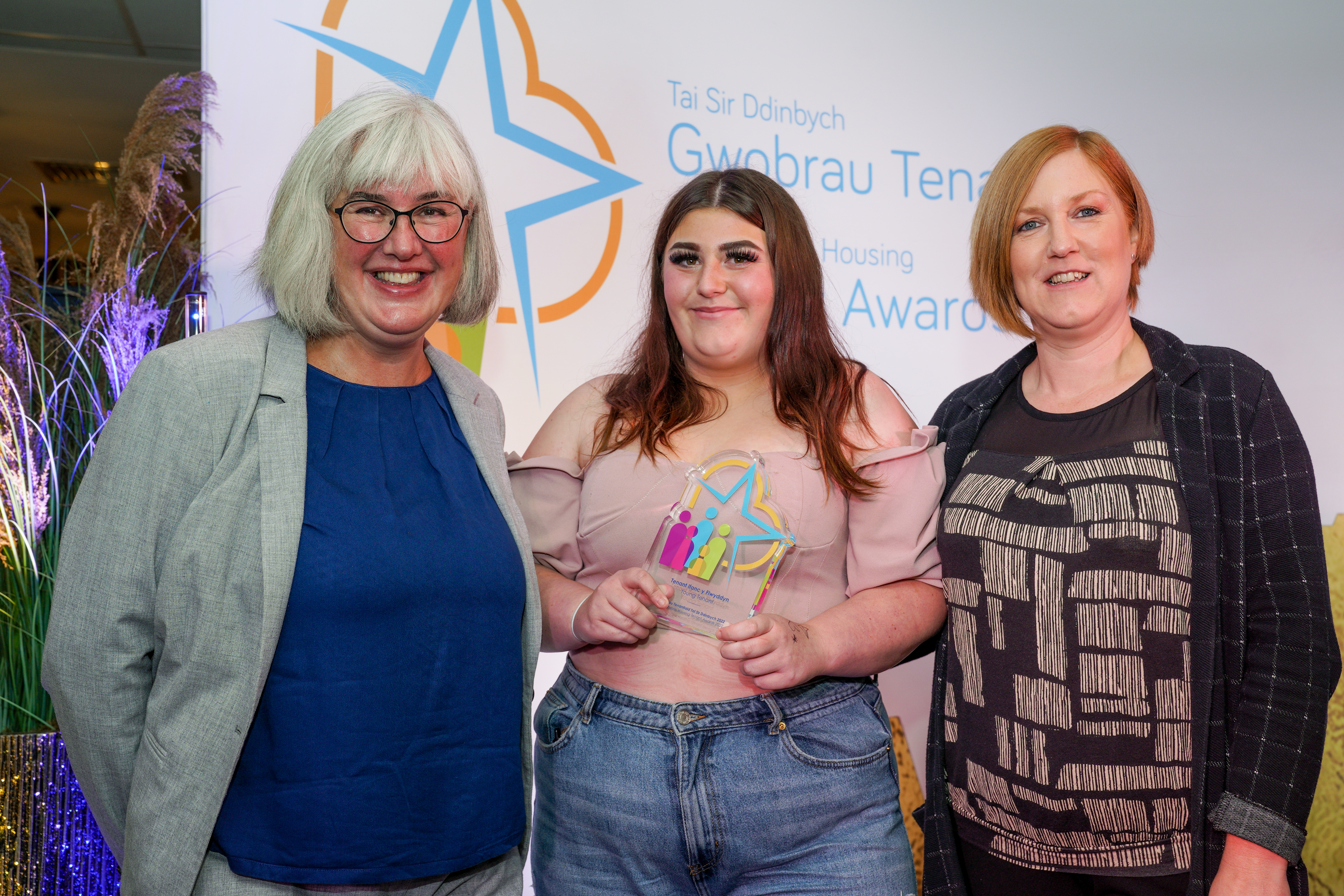 Three women stood together and the woman in the middle is holding a colourful trophy.