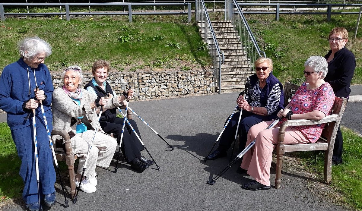 A small group of older women outside, holding walking sticks and taking part in armchair exercises.
