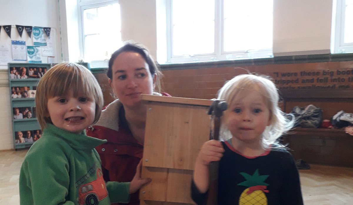 Two young children and a female adult standing in a hall, holding a bird box that they have built as part of the Nature for Health project.